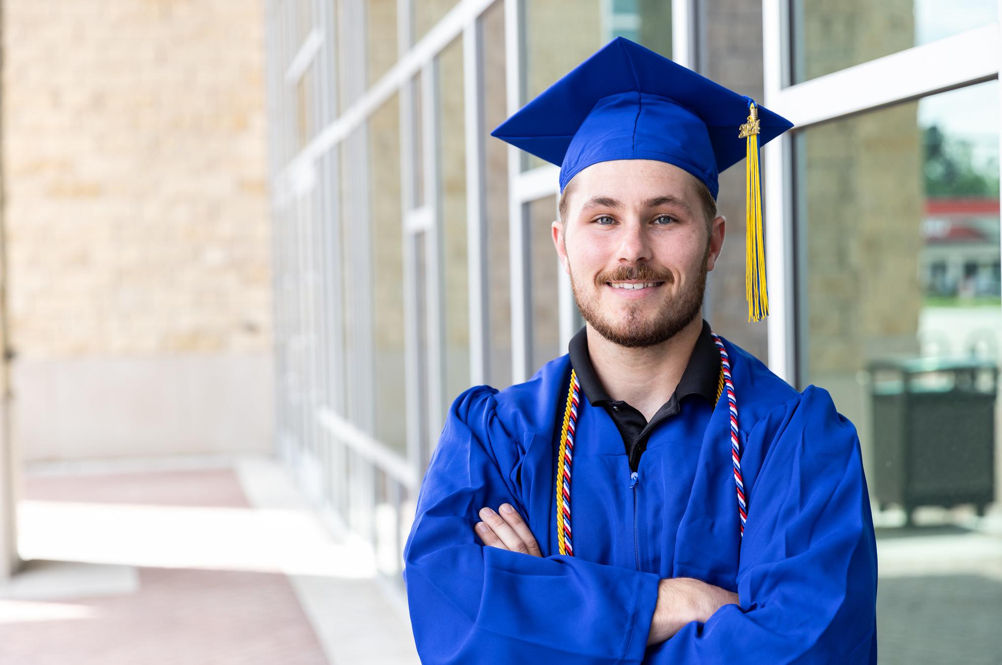 man standing in blue cap and gown outside of a building with a lot of windows