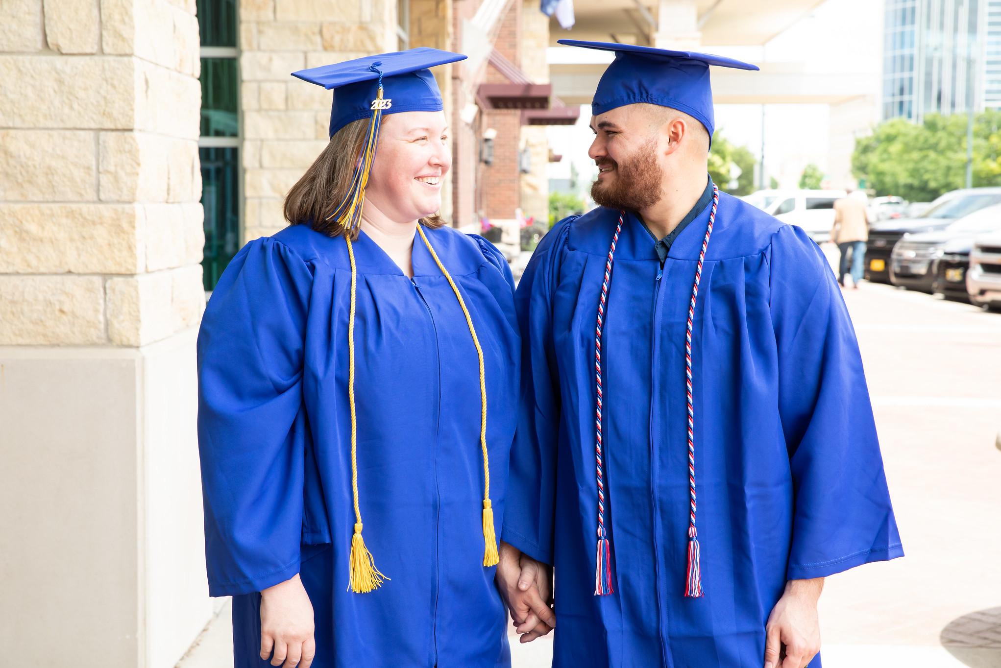 two graduates in caps and gowns smiling and looking at each other while holding hands