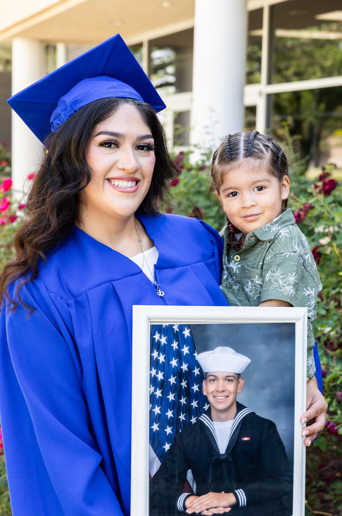 woman in cap and gown holding her son and a picture of her husband in navy uniform