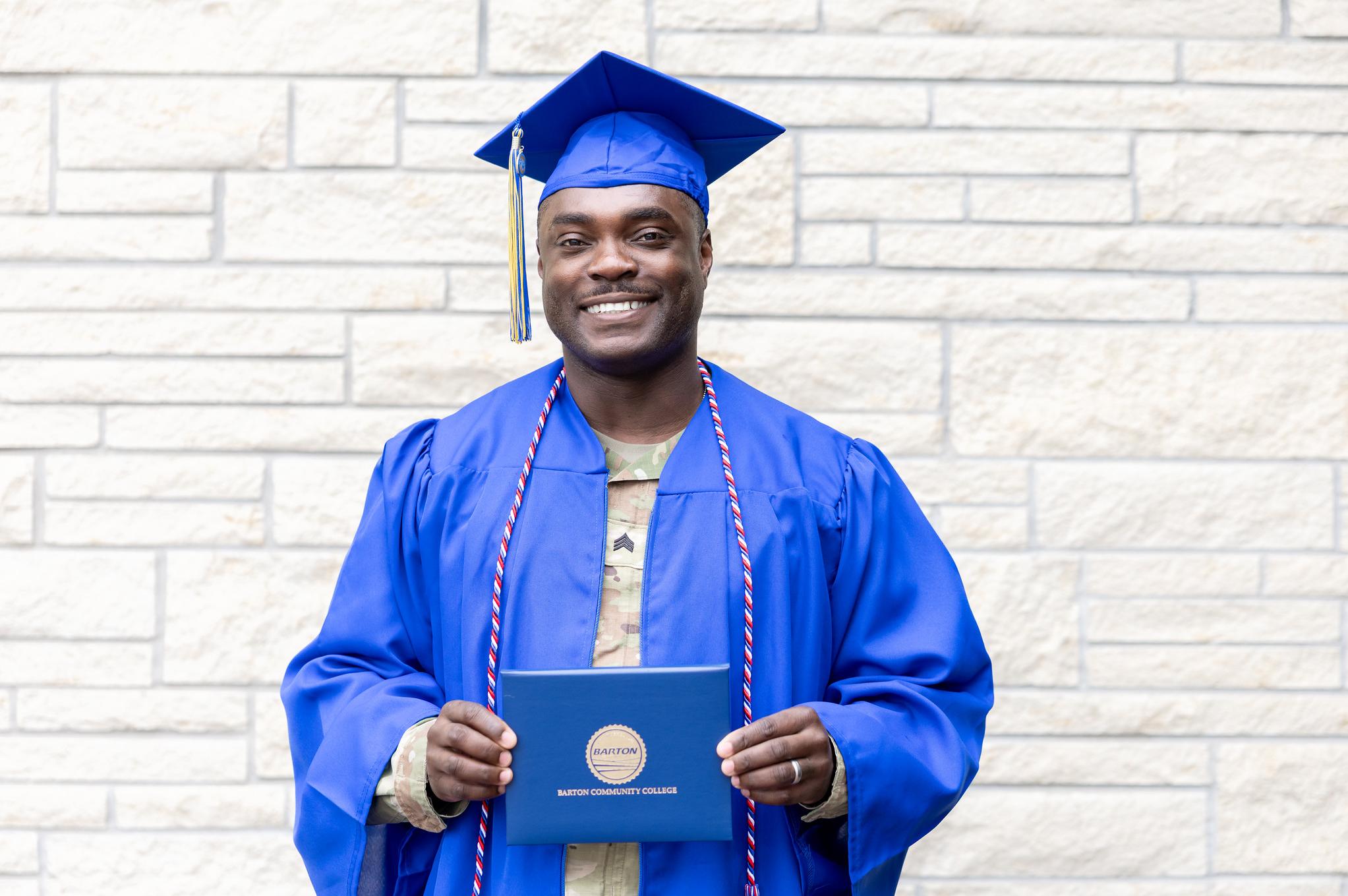 graduate in cap and gown and army fatigues smiling and holding diploma