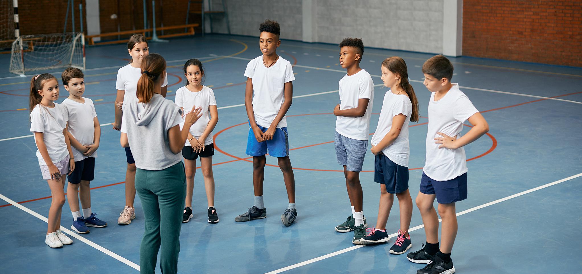 A group of children in a gymnasium listening to a teacher.