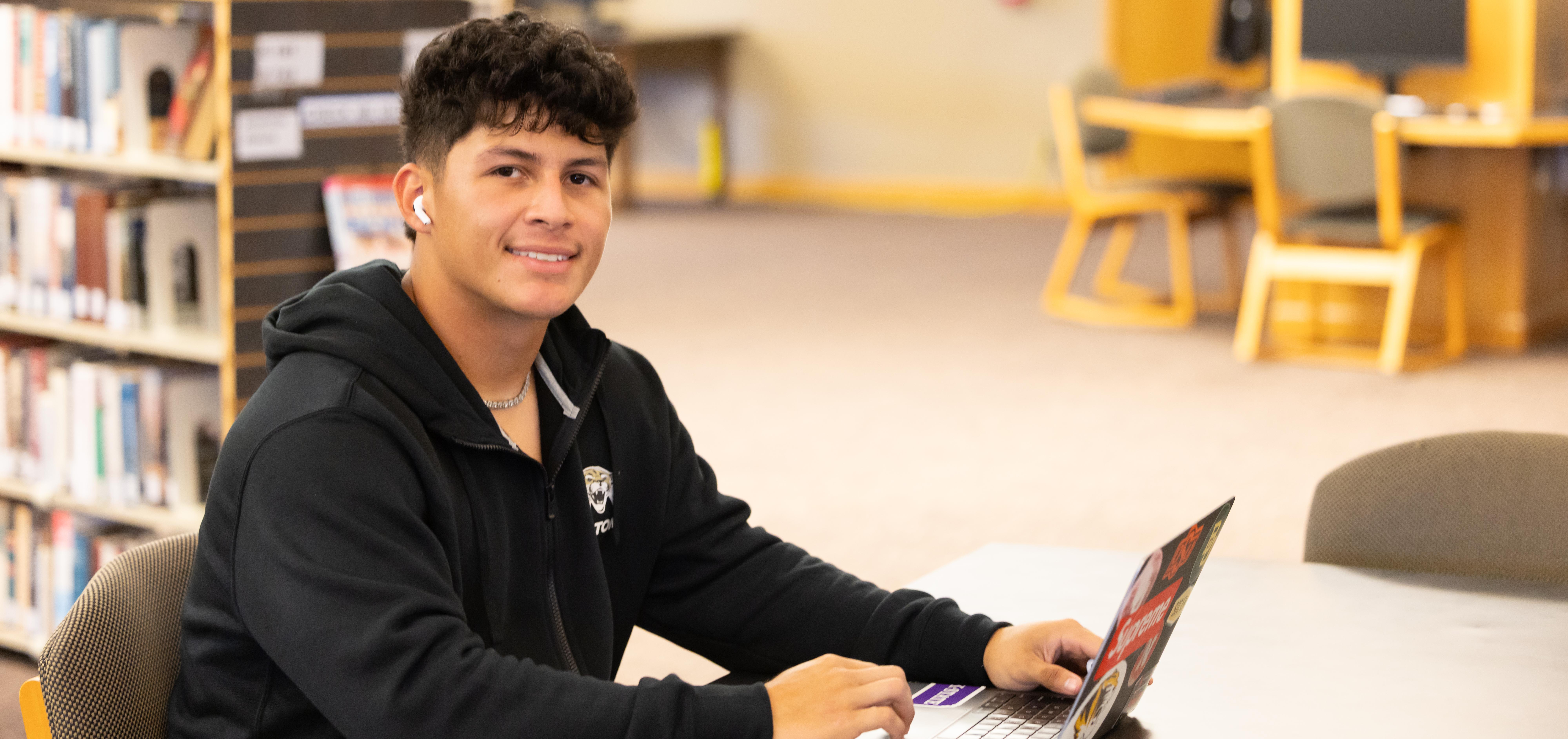 A student works on a laptop.  He wears an earbud in his ear and is smiling at the camera.
